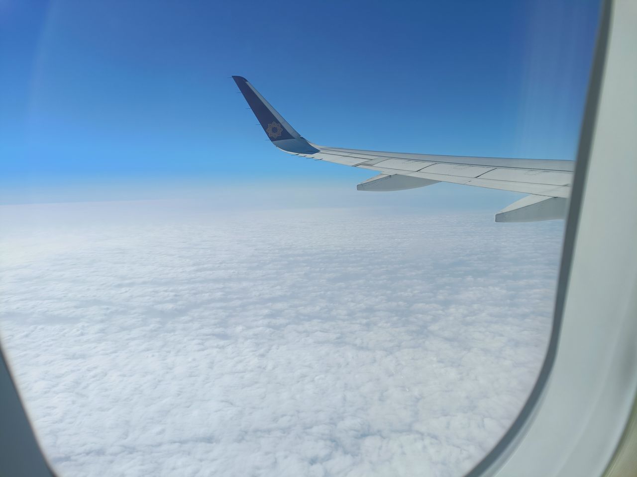 View of an airplane wing above clouds under a clear blue sky, captured from a window seat.